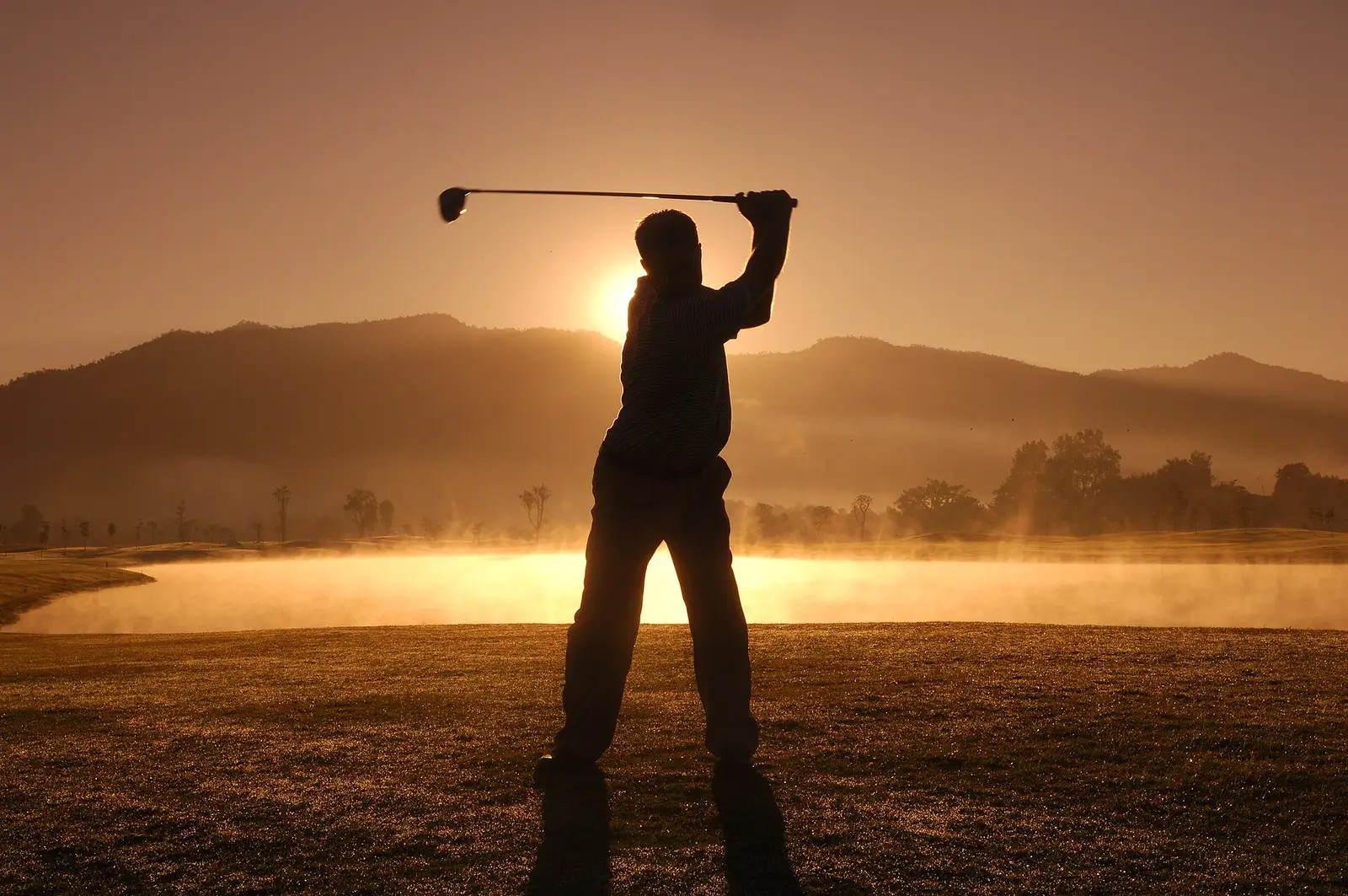 A golfer mid-swing on the practice ground, silhouetted against the setting sun